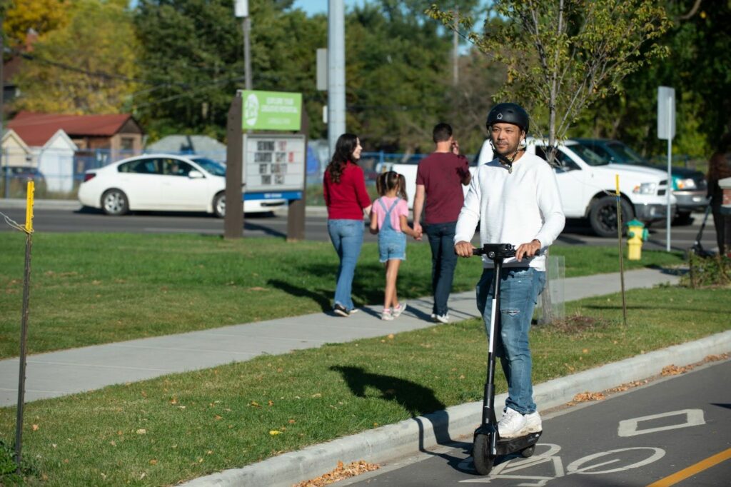 Man in a white shirt and jeans using an e-scooter in a protected bike line on a sunny day.