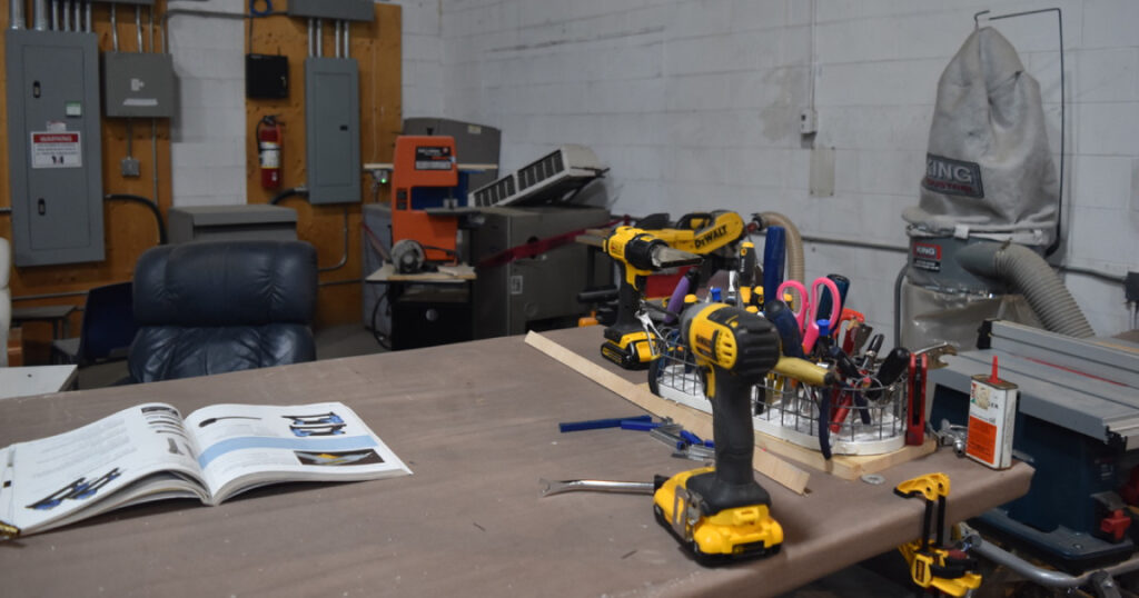 An open book sits in the middle of work bench with a bunch of tools, a basket of scissors, felt pens and other implements, sitting near the right edge of the table.