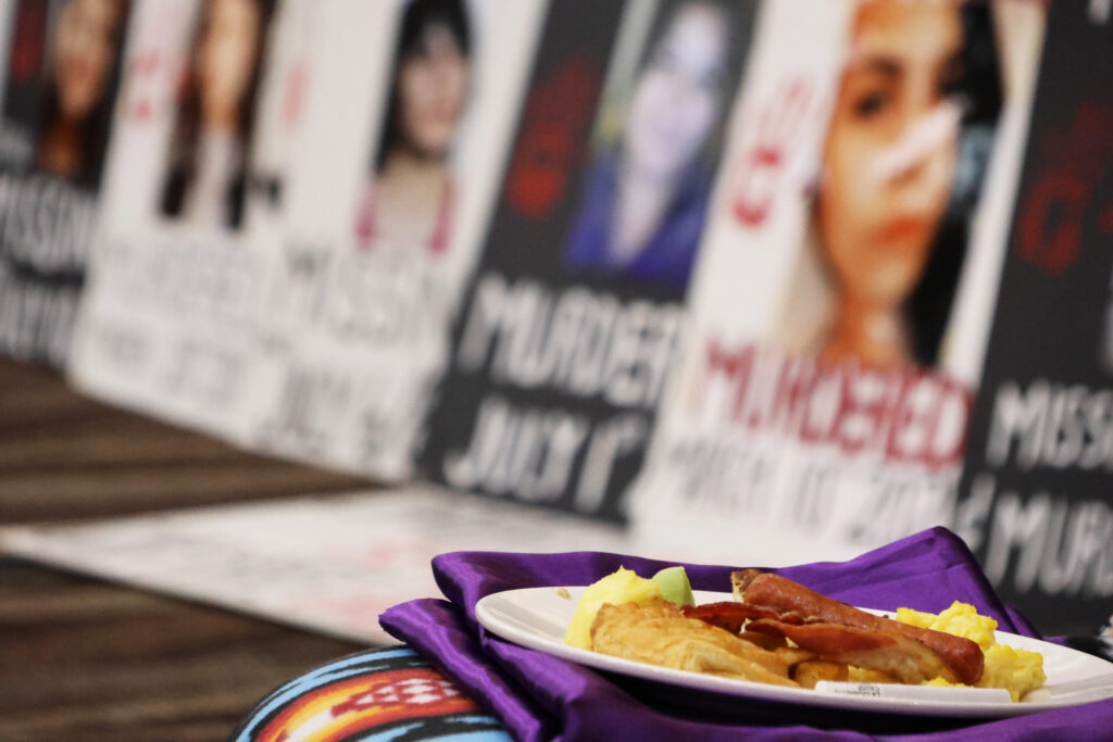 A spirit plate is laid out at the MMIR Gathering event with posters of Missing and Murdered Indigenous Relative posters in the background.