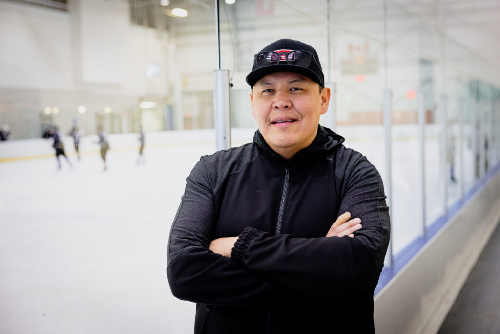 An Indigenous man stands with his arms crossed next to an ice rink with some players in the distance.