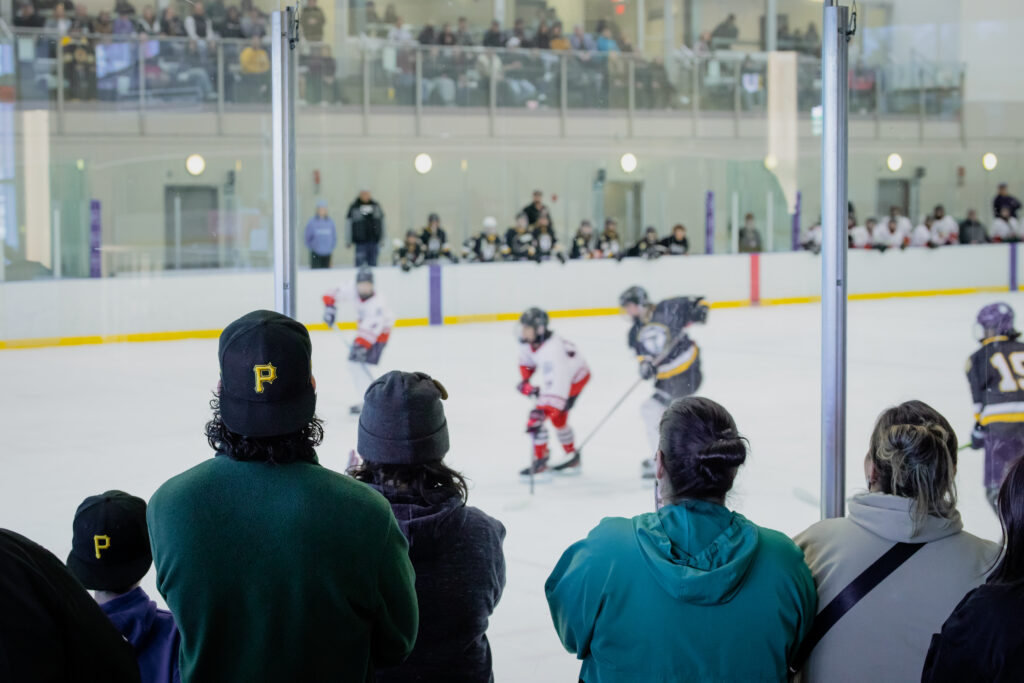 People wearing baseball caps and hair buns stand with their backs to the camera as they look through the glass and watch young Indigenous hockey players on the ice.
