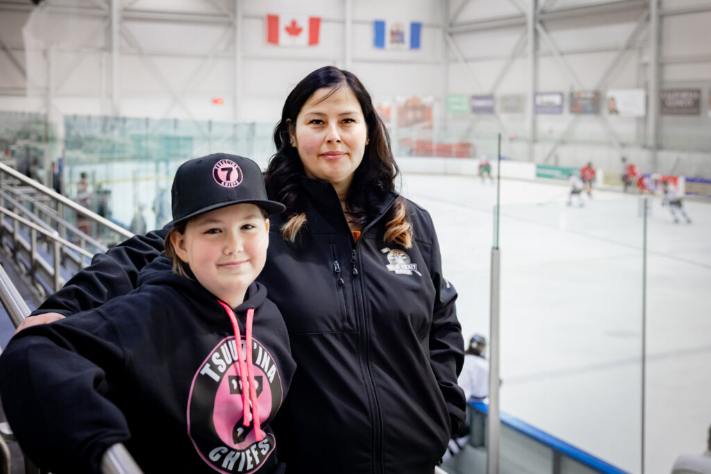 An Indigenous mom and her daughter sit in the stands at a hockey game.