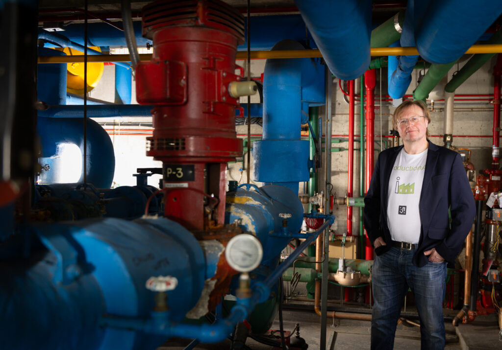 A man stands next to some blue, red and green pipes on the wall, the ceiling and beside him.