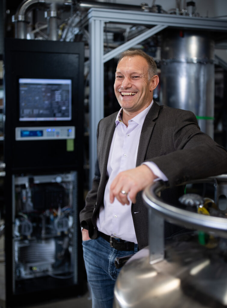 A man smiles as he stands in front of a dilution refrigerator.